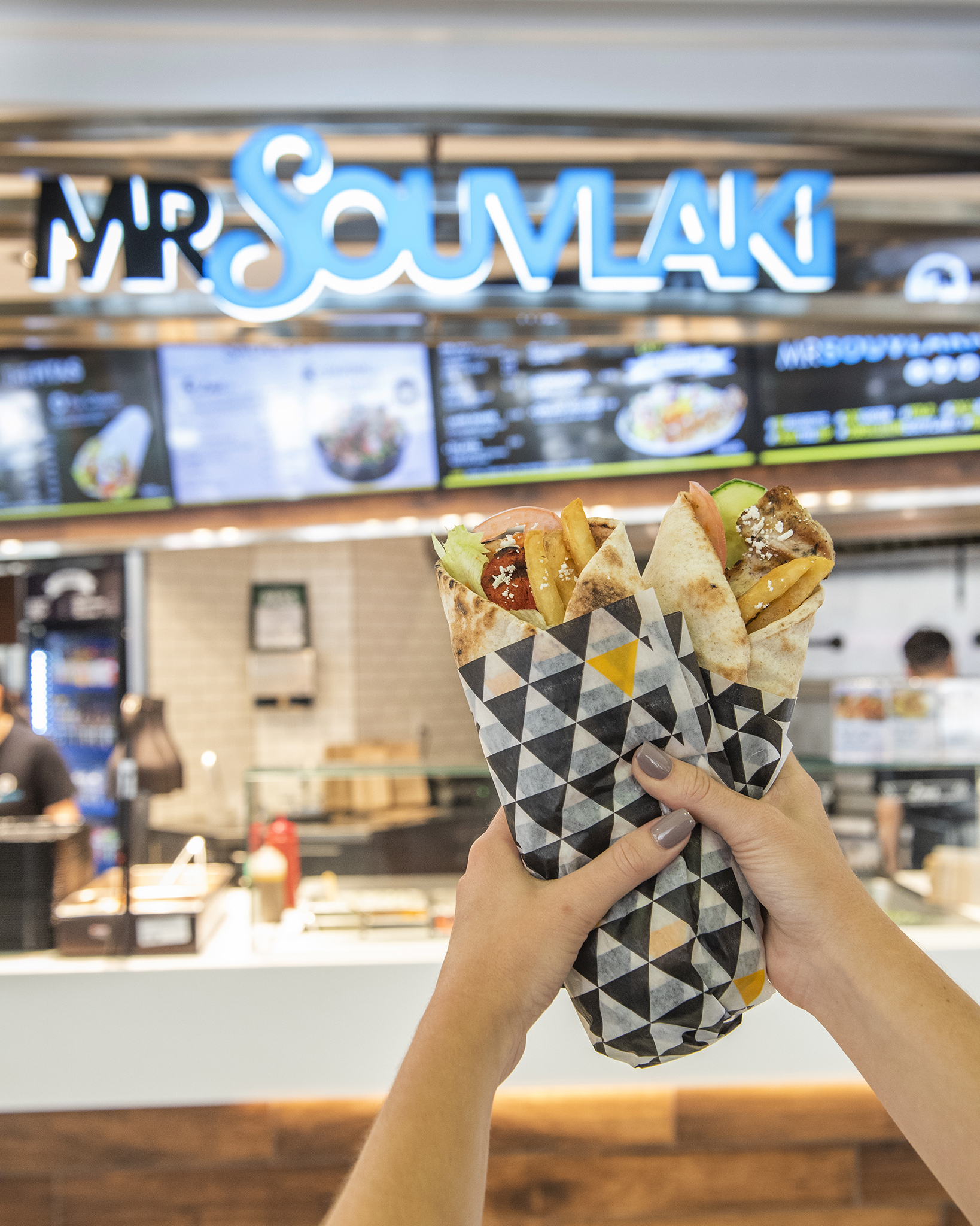 Hands holding Greek wraps in front of a Mr. Souvlaki restaurant sign.