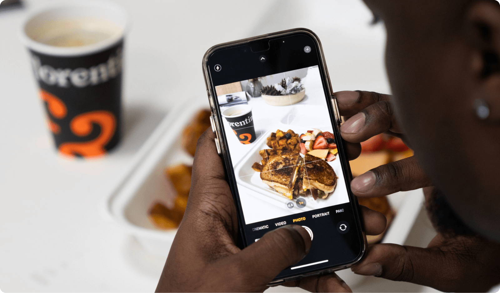 Person holding a smartphone, taking a photo of a meal with French toast, potatoes, and fruit, with a coffee cup in the background.