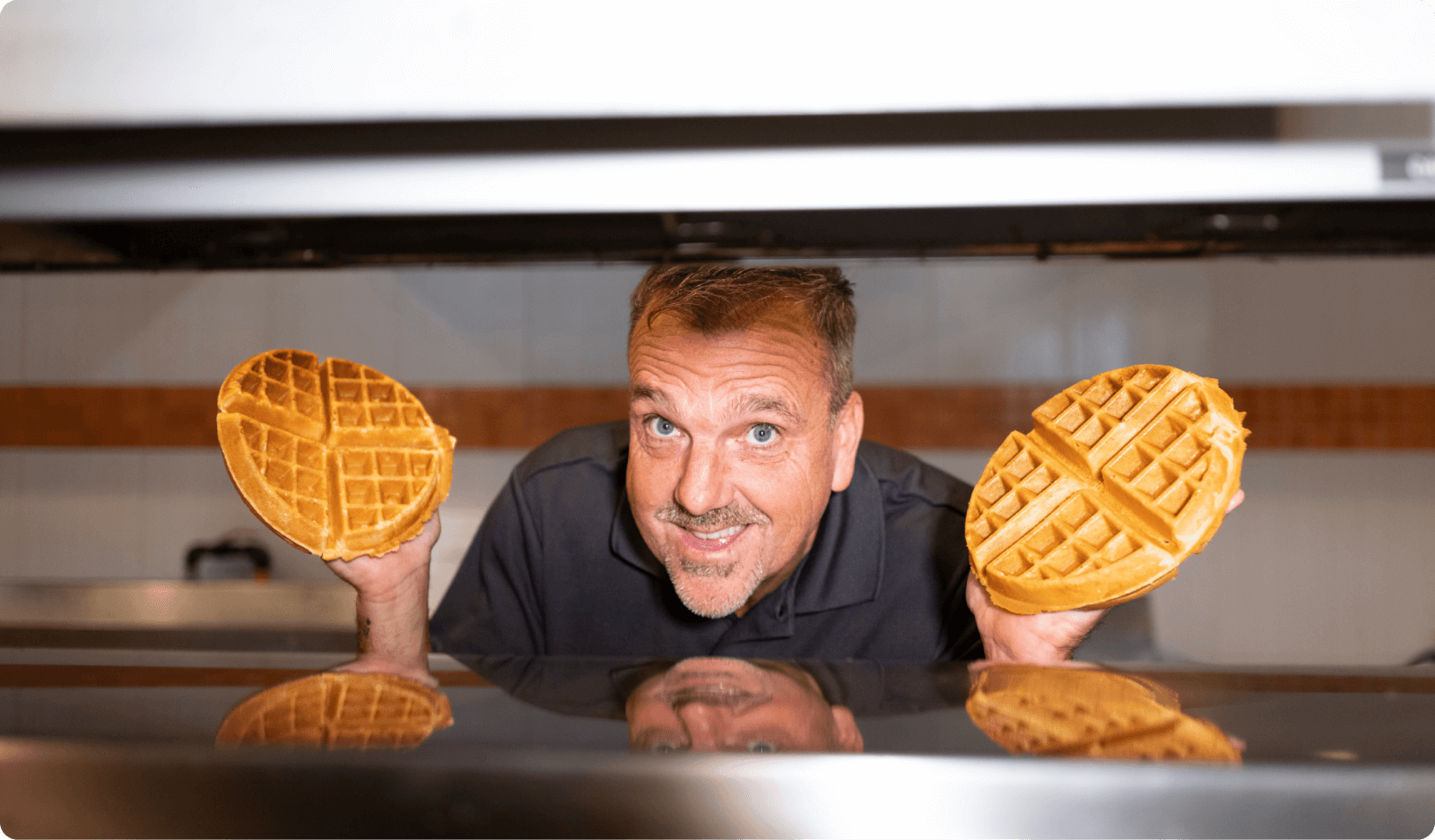 Man smiling while holding two waffles, one in each hand, with a kitchen backdrop.