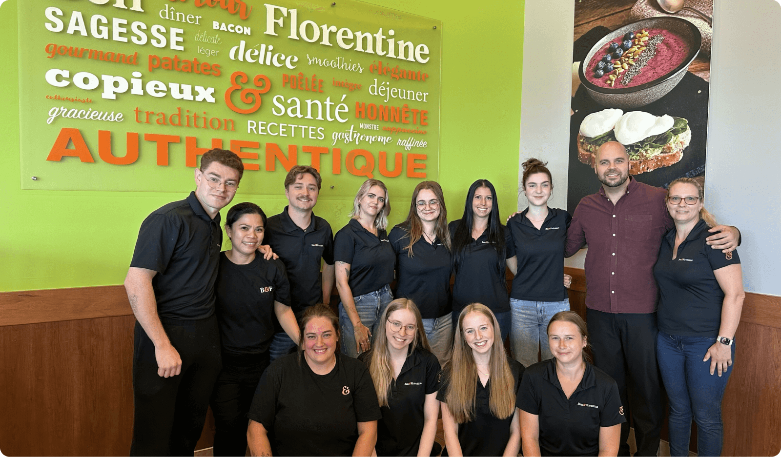 Group photo of a restaurant staff in black shirts standing in front of a colorful wall with menu items, smiling and posing together.