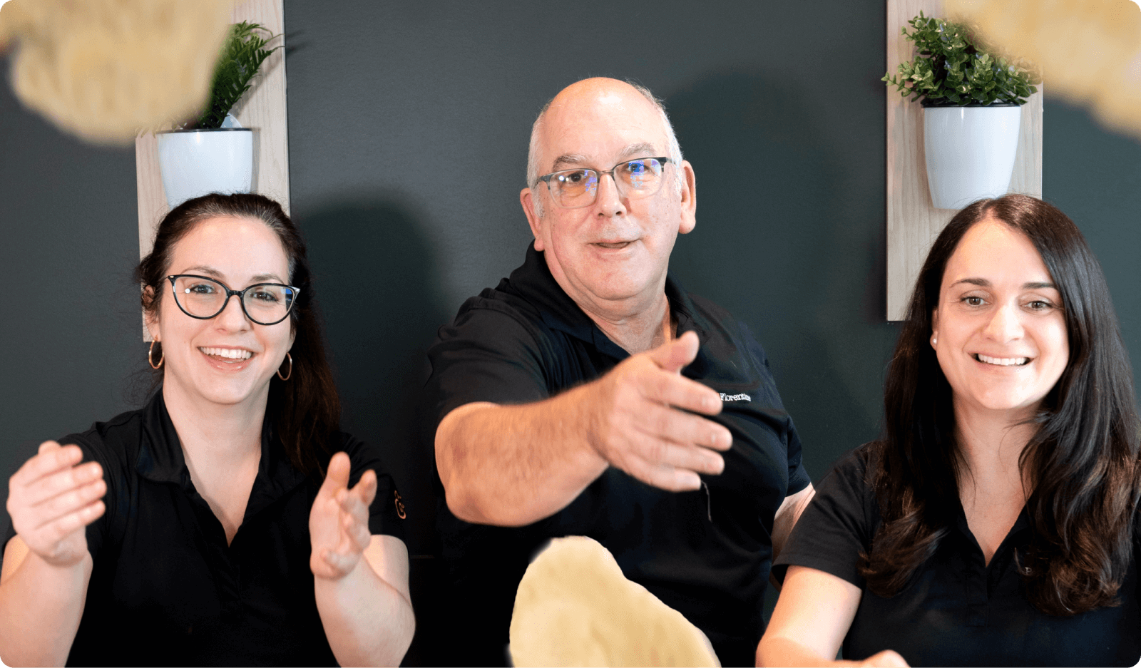 Three people in black shirts sitting together, with one man reaching out and two women smiling, against a neutral background.