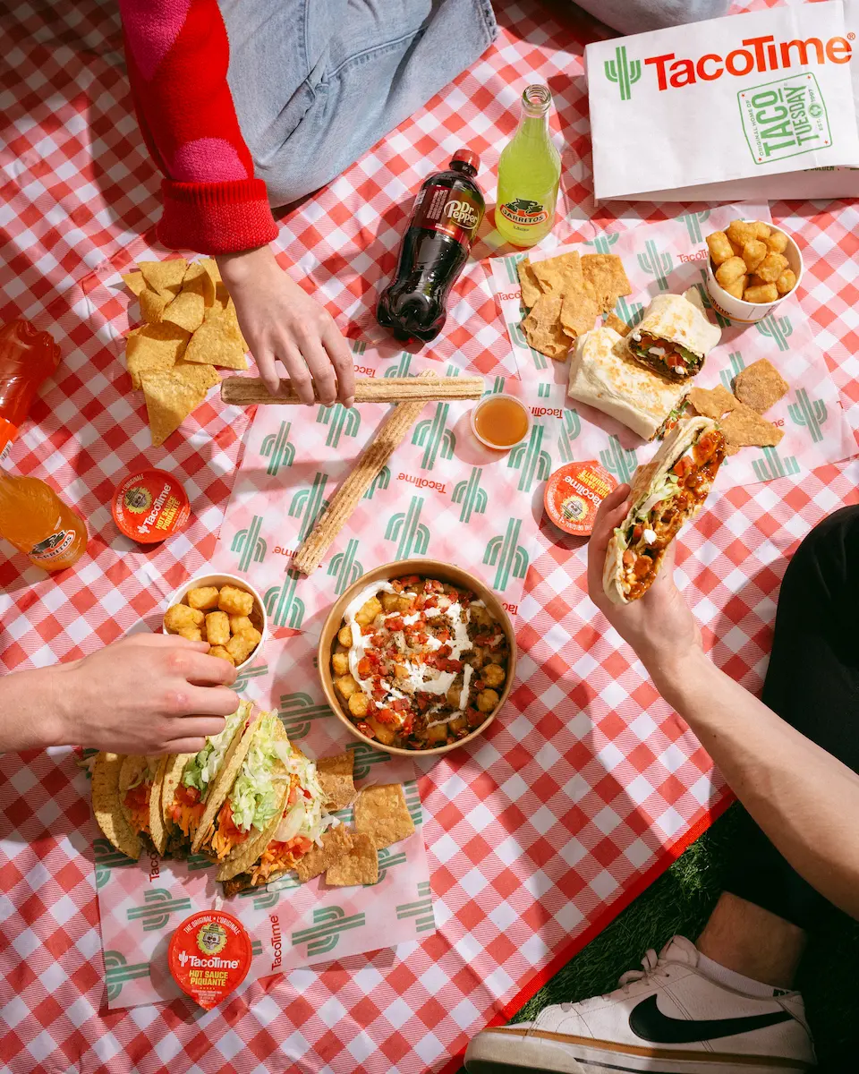 Overhead view of a TacoTime picnic on a red checkered blanket with tacos, burritos, churros, tater tots, and drinks shared among friends.